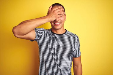 Genç yakışıklı adam sarı izole arka plan üzerinde çizgili t-shirt giyen gülümseyerek ve sürpriz için gözleri kaplayan yüzünde el ile gülüyor. Kör kavramı.
