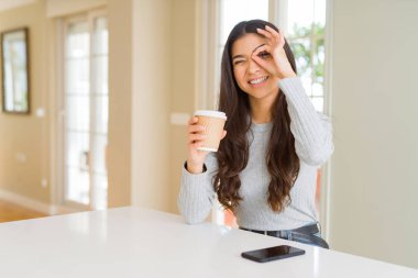Mutlu yüzü ile evde bir fincan kahve içen genç kadın parmaklarıile bakarak göz üzerinde el ile ok işareti yapıyor gülümseyerek