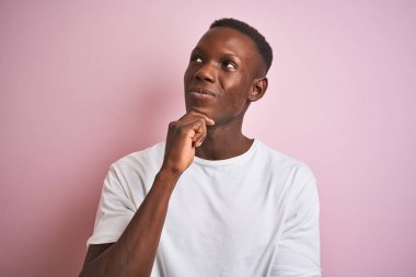 Izole pembe arka plan üzerinde duran beyaz t-shirt giyen Afro-Amerikan adam soru hakkında ciddi yüz düşünme, çok karışık bir fikir