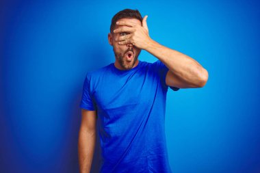 Genç yakışıklı adam mavi izole arka plan üzerinde rahat t-shirt giyen şok yüz ve el ile gözleri kapsayan gözetleme, utanmış ifade ile parmaklarından bakarak.