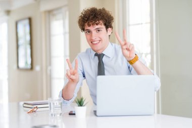 Ofiste bilgisayar dizüstü bilgisayarıyla çalışan genç iş adamı gülümseyerek kameraya bakarak parmaklarını göstererek zafer işareti yapıyor. İki numara.