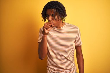 Dreadlocks çizgili t-shirt izole sarı arka plan üzerinde ayakta giyen Afro adam rahatsız hissediyorum ve soğuk veya bronşit için belirti olarak öksürük. Sağlık kavramı.