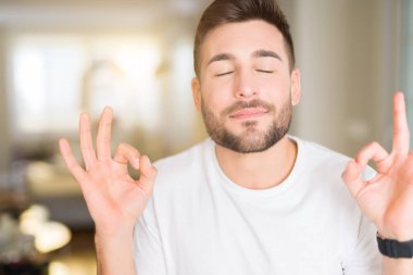 Casual beyaz t-shirt evde dinlenmek giydiği ve gözleri ile gülümseyen yakışıklı delikanlı parmak ile meditasyon jest yapıyor kapalı. Yoga kavramı.
