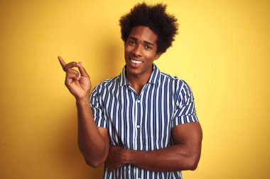 Afro saçlı amerikalı adam yüzünde büyük bir gülümseme ile izole sarı arka plan üzerinde duran çizgili gömlek giyen, kameraya bakarak yan el ve parmak ile işaret.
