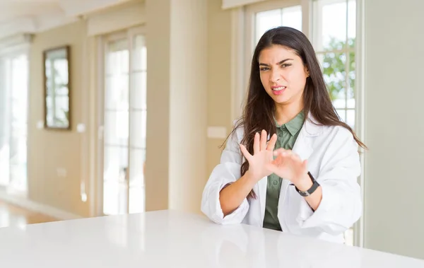 Young woman wearing medical coat at the clinic as therapist or doctor disgusted expression, displeased and fearful doing disgust face because aversion reaction. With hands raised. Annoying concept.