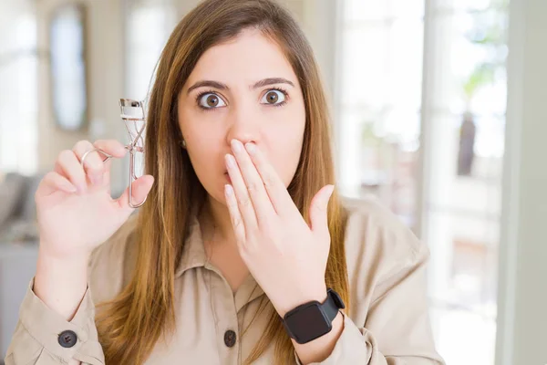 Beautiful young woman curling eyelashes using eyelash curler cover mouth with hand shocked with shame for mistake, expression of fear, scared in silence, secret concept