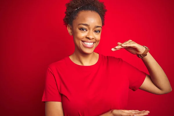 Young beautiful african american woman with afro hair over isolated red background gesturing with hands showing big and large size sign, measure symbol. Smiling looking at the camera. Measuring concept.