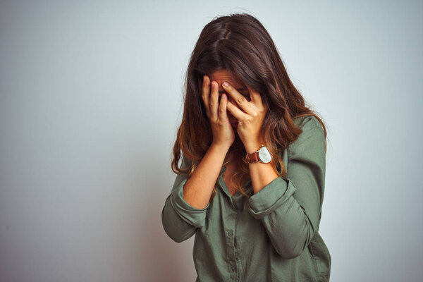 Young beautiful woman wearing green shirt standing over grey isolated background with sad expression covering face with hands while crying. Depression concept.