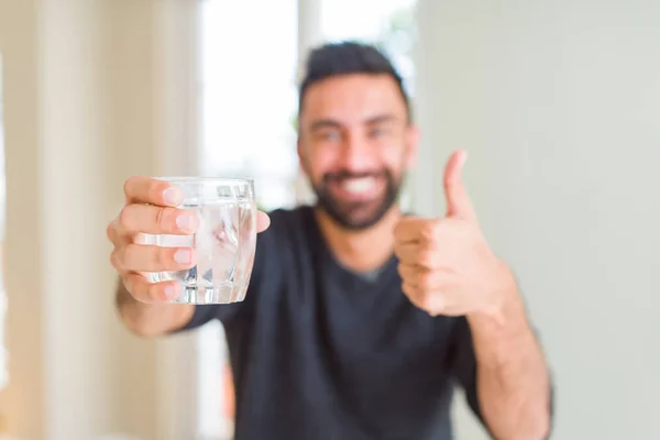 Handsome hispanic man drinking a fresh glass of water happy with big smile doing ok sign, thumb up with fingers, excellent sign