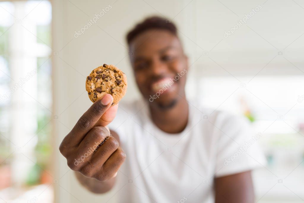 Young african american man eating chocolate chips cookies with a happy face standing and smiling with a confident smile showing teeth