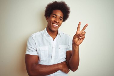 Afro saçlı afro saçlı adam izole beyaz arka plan üzerinde duran mutlu yüz zafer işareti yapıyor kameraya göz kırparak gülümseyerek gömlek giyiyor. İki numara.