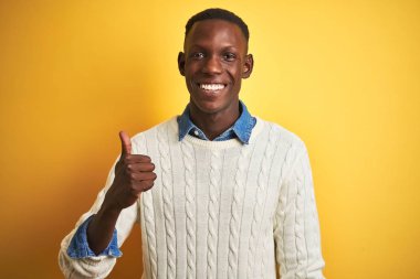 Denim gömlek ve izole sarı arka plan üzerinde beyaz kazak giyen Afro-Amerikan adam el ile jest kadar mutlu başparmak yapıyor. Başarı gösteren kameraya bakarak ifadeyi onaylama.