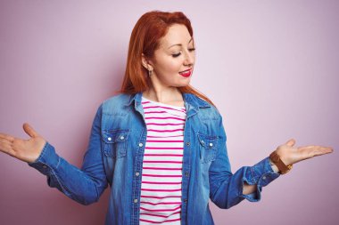 Denim gömlek giyen güzel kızıl saçlı kadın ve izole pembe arka plan üzerinde çizgili t-shirt gülümseyerek her iki elaçık avuç içi gösteren, sunan ve reklam karşılaştırma ve denge