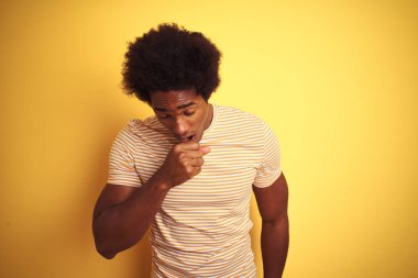 Afro saçlı Amerikalı adam izole sarı arka plan üzerinde ayakta çizgili t-shirt giyen rahatsız hissetme ve soğuk veya bronşit için bir belirti olarak öksürük. Sağlık kavramı.