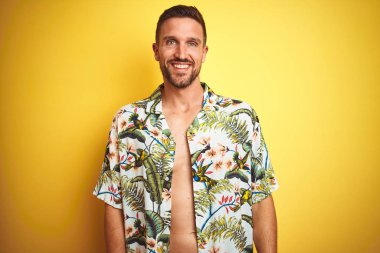 Yakışıklı adam yüzünde mutlu ve serin bir gülümseme ile sarı izole arka plan üzerinde yaz hawaii çiçek gömlek giyiyor. Şanslı kişi.