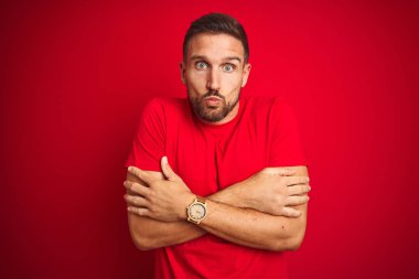 Genç yakışıklı adam yüzünde üzgün ve şok ifade ile kış soğuk için sallayarak ve donma kırmızı izole arka plan üzerinde rahat t-shirt giyiyor