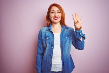 Youg güzel kızıl saçlı kadın izole pembe arka plan üzerinde duran denim gömlek giyen gösteren ve emin ve mutlu gülümserken parmakları ile dört numara işaret.