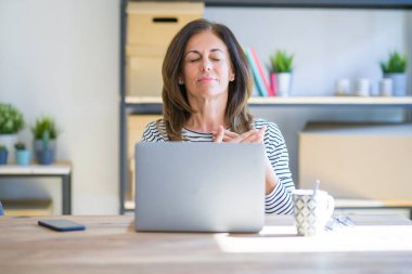 Orta yaş yaşlı kadın evde masada oturan kapalı gözleri ve yüzünde minnettar jest göğüs elleri ile gülümseyen bilgisayar dizüstü bilgisayar kullanarak çalışıyor. Sağlık kavramı.