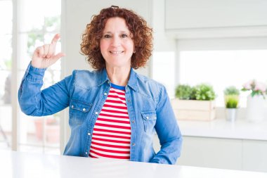 Kıvırcık saçlı orta yaş yaşlı kadın evde kot ceket giyen gülümseyen ve emin el parmakları görünümlü ve kamera ile küçük boy işareti yapıyor gesturing. Ölçü kavramı.