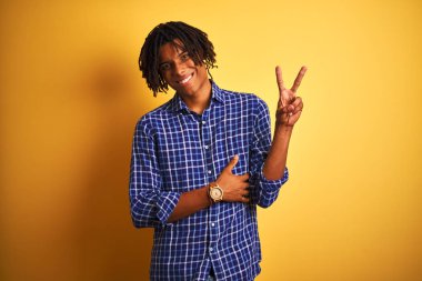 Rahat gömlek giyen dreadlocks ile Afro adam mutlu yüz zafer işareti yapıyor kameraya göz kırparak ile gülümseyerek izole sarı arka plan üzerinde duran rahat gömlek giyiyor. İki numara.