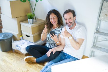 Orta yaş yaşlı çift ok işareti yapıyor büyük bir gülümseme ile mutlu akıllı telefon kullanarak yeni bir eve hareket, parmakları ile başparmak, mükemmel işareti