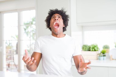 Genç afro-amerikan adam evde oturan rahat beyaz t-shirt giyen kızgın ve deli çığlık sinirli ve öfkeli, öfke ile bağırıyor. Öfke ve agresif kavram.