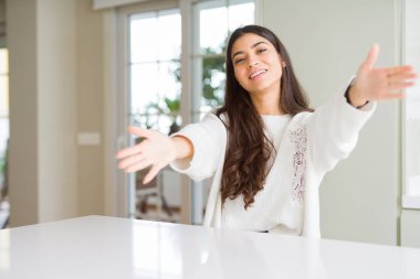 Genç güzel kadın evde beyaz masada kameraya bakarak sarılmak için kollarını açarak gülümsüyor. Mutluluğu kucaklayan neşeli ifade.