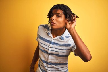 Izole sarı arka plan üzerinde çizgili gömlek giyen dreadlocks ile Afro Amerikan adam kulak üzerinde el ile gülümseyerek söylenti veya dedikodu bir işitme dinlerken. Sağırlık kavramı.