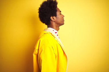 Afro saçlı afro saçlı afro-amerikan adam yan bakarak izole sarı arka plan üzerinde duran yağmurluk giyen, kendine güvenen gülümseme ile doğal yüz ile profil poz dinlenmek.