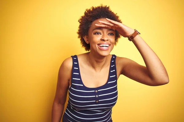 Beauitul african american woman wearing summer t-shirt over isolated yellow background very happy and smiling looking far away with hand over head. Searching concept.