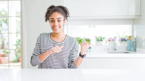 Beautiful african american woman with afro hair wearing casual striped sweater Pointing to the back behind with hand and thumbs up, smiling confident