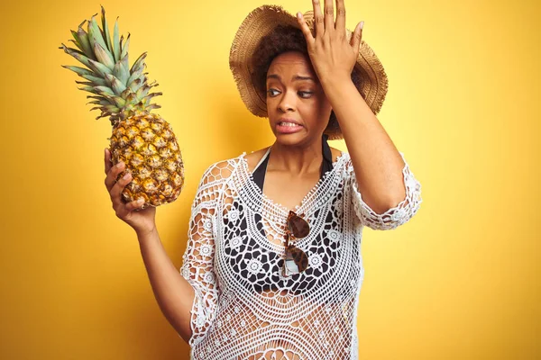 Young african american woman with afro hair holding pineaple over yellow isolated background stressed with hand on head, shocked with shame and surprise face, angry and frustrated. Fear and upset for mistake.