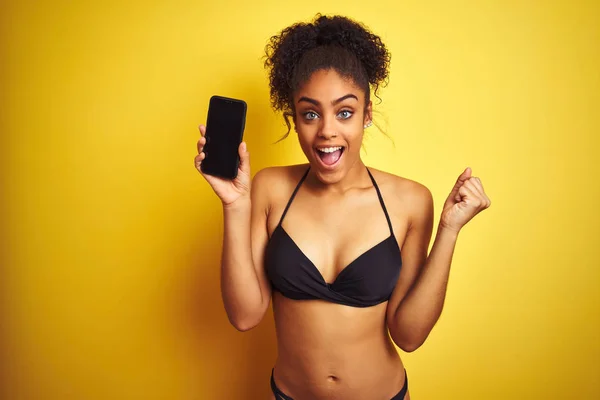 African american woman wearing bikini showing smartphone over isolated yellow background screaming proud and celebrating victory and success very excited, cheering emotion