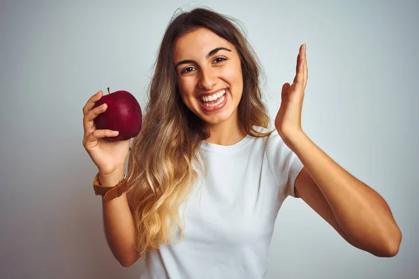 Young beautiful woman eating red apple over grey isolated background very happy and excited, winner expression celebrating victory screaming with big smile and raised hands