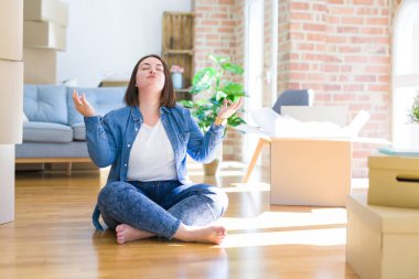 Genç artı boyutu kadın karton kutular etrafında yeni bir eve hareket yerde oturan dinlenmek ve gözleri parmakları ile meditasyon jest yapıyor kapalı gülümseyerek. Yoga konsepti.