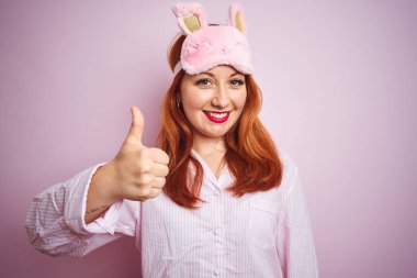 Genç güzel kızıl saçlı kadın pembe izole arka plan üzerinde pijama giyen el ile jest kadar mutlu başparmak yapıyor. Başarı göstererek kameraya bakan ifadeyi onaylama.