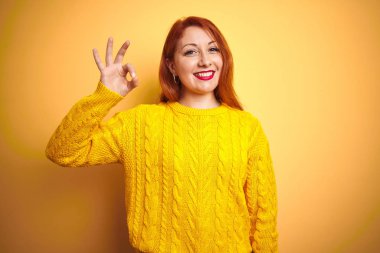 Güzel kızıl saçlı kadın izole sarı arka plan üzerinde duran kış kazak giyen olumlu el ve parmakları ile ok işareti yapıyor gülümseyerek. Başarılı ifade.