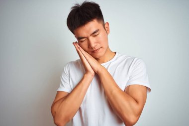 Genç asyalı Çinli adam izole beyaz arka plan üzerinde duran t-shirt giyen yorgun rüya ve kapalı gözlerle gülümseyerek birlikte elleri ile poz.