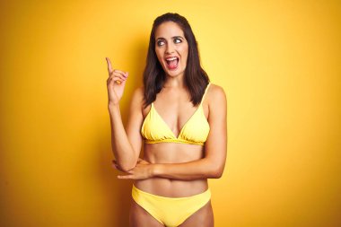 Güzel kadın yüzünde büyük bir gülümseme ile izole sarı arka plan üzerinde yaz aylarında sarı bikini giyen, kameraya bakarak yan el ve parmak ile işaret.