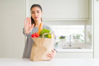 Açık el ile poşet dolu poşet tutan genç kadın ciddi ve kendinden emin ifade, savunma jesti ile dur işareti yapıyor