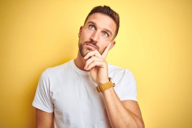 Genç yakışıklı adam soru, dalgın ifade hakkında düşünme çene üzerinde el ile sarı izole arka plan üzerinde rahat beyaz t-shirt giyiyor. Düşünceli bir yüzle gülümsüyor. Şüphe kavramı.