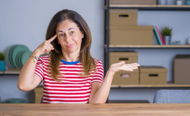 Orta yaş yaşlı kadın evde masada oturan şaşkın ve açık avuç içi ile rahatsız kopya alanı gösteren ve alnına parmak işaret. Bir düşün..