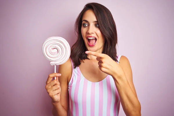 Young beautiful woman eating sweet candy over pink isolated background very happy pointing with hand and finger