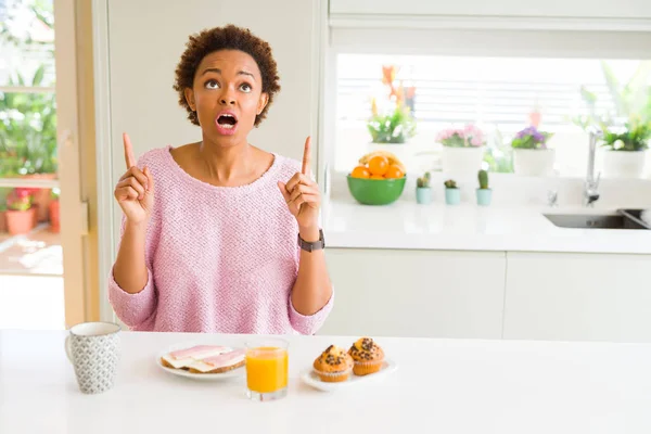 年轻的非洲裔美国妇女吃休息快 在早上在家里惊讶和惊讶抬头 用手指和抬起手臂 — 图库照片