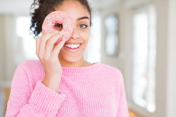 幸せな顔で甘いピンクのドーナツを食べる若いアフリカ系アメリカ人の女の子は 歯を見せる自信のある笑顔で立って微笑んでいます — ストック写真