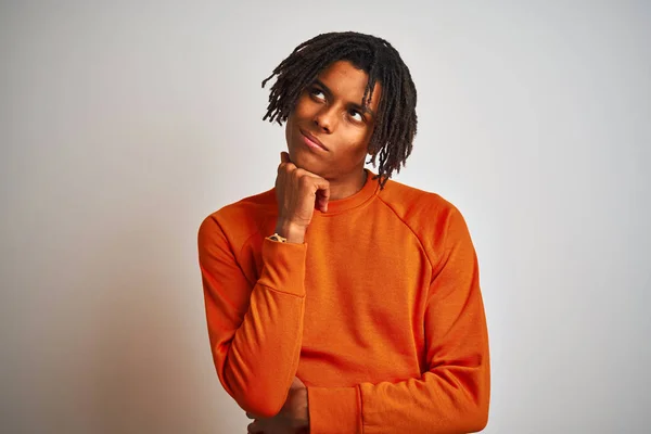 Afro american man with dreadlocks wearing orange sweater over isolated white background with hand on chin thinking about question, pensive expression. Smiling with thoughtful face. Doubt concept.