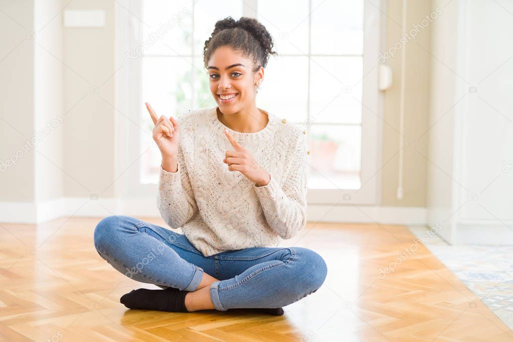 Beautiful young african american woman with afro hair sitting on the floor smiling and looking at the camera pointing with two hands and fingers to the side.