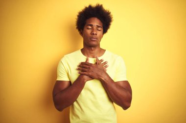 Afro saçlı afro saçlı adam izole sarı arka plan üzerinde duran t-shirt giyen kapalı gözleri ve yüzünde minnettar jest göğüs elleri ile gülümseyerek. Sağlık kavramı.