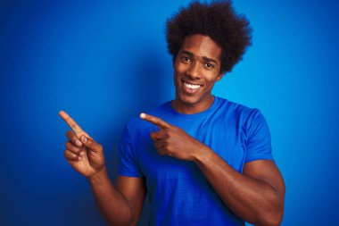 Afro saçlı Afro saçlı adam izole mavi arka plan üzerinde duran t-shirt giyen gülümseyerek ve yan iki el ve parmakları ile işaret kamera bakıyor.