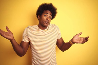 Afro saçlı Amerikalı adam çizgili t-shirt izole sarı arka plan üzerinde duran giyen habersiz ve kolları ve elleri ile karışık ifade kaldırdı. Şüphe kavramı.